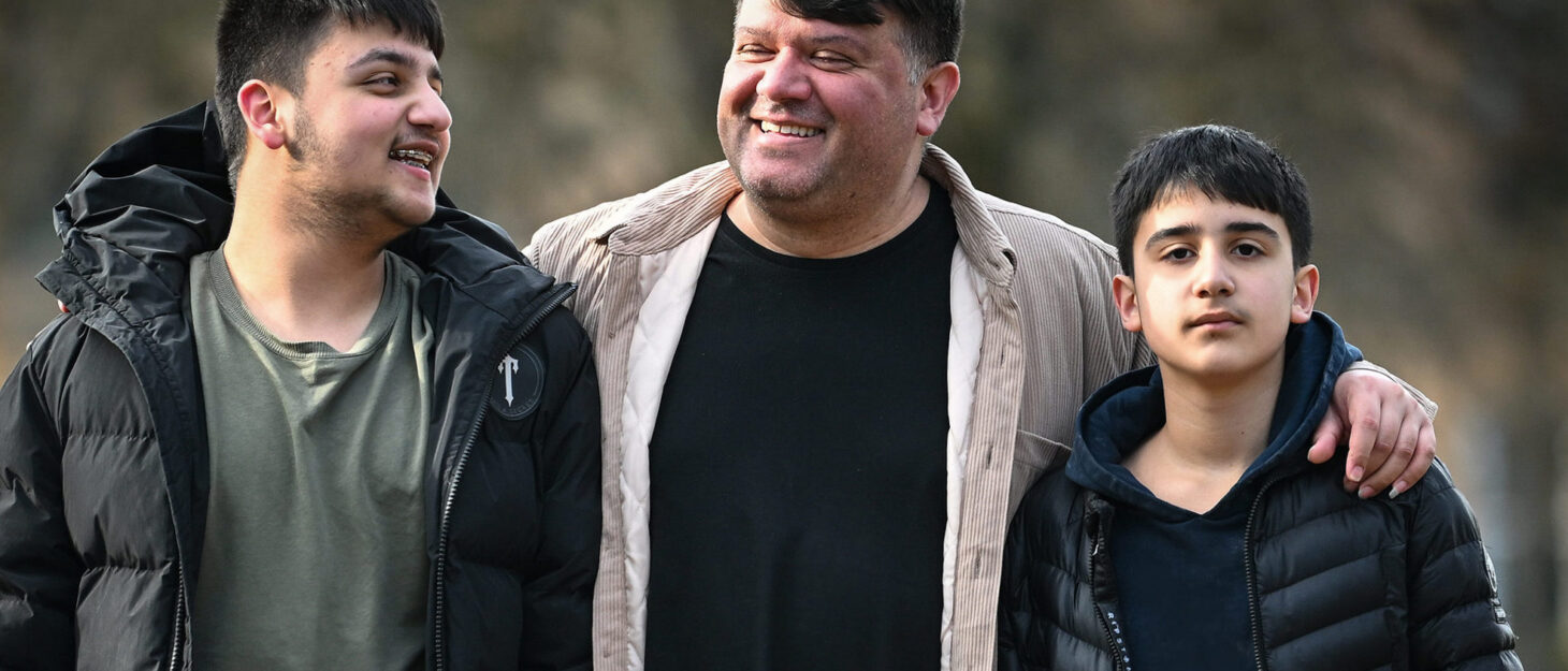 A dad and his two sons outdoors in the sunshine, dad's arms wrapped around the boys.