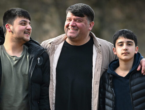 A dad and his two sons outdoors in the sunshine, dad's arms wrapped around the boys.