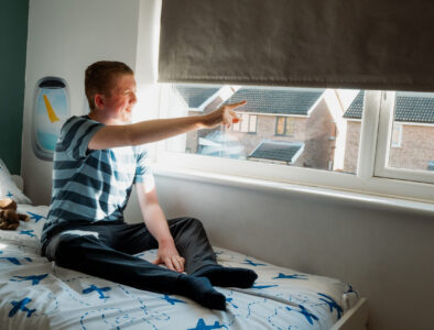 A teenage boy sits on his bed, pointing out of his window to the sky. His bedding has aeroplanes on it.