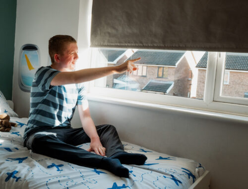 A teenage boy sits on his bed, pointing out of his window to the sky. His bedding has aeroplanes on it.