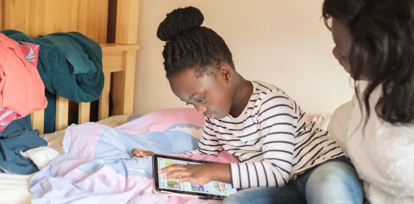A mum and her daughter sit on the bed playing on the ipad together