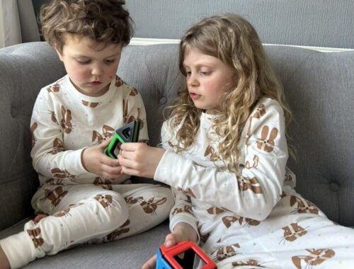Two children sit on a sofa together in matching pjs, playing with a Polydron magnetic building set.