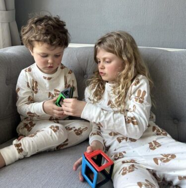 Two children sit on a sofa together in matching pjs, playing with a Polydron magnetic building set.