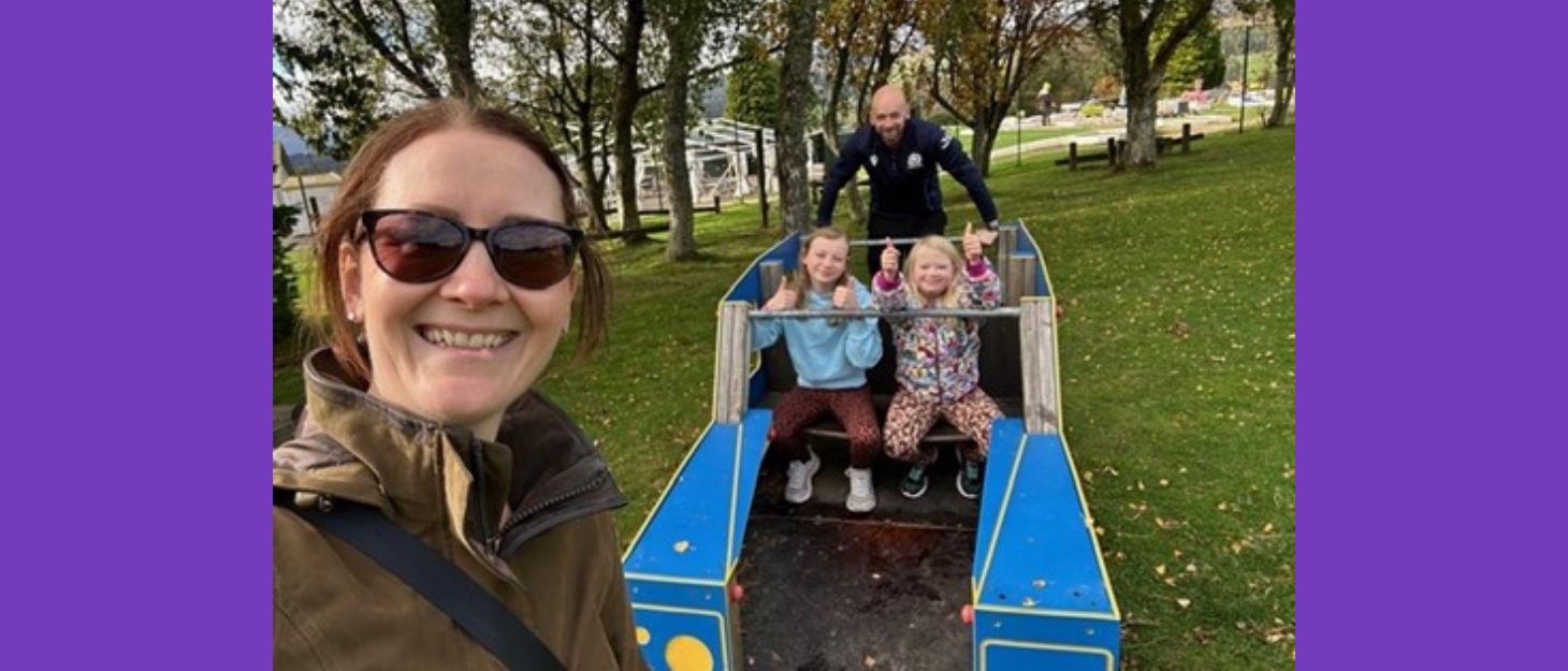 A family of two adults and two children play together outdoor on play equipment.