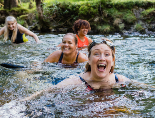 A diverse group of women wild swim in a lake, and are smiling joyfully