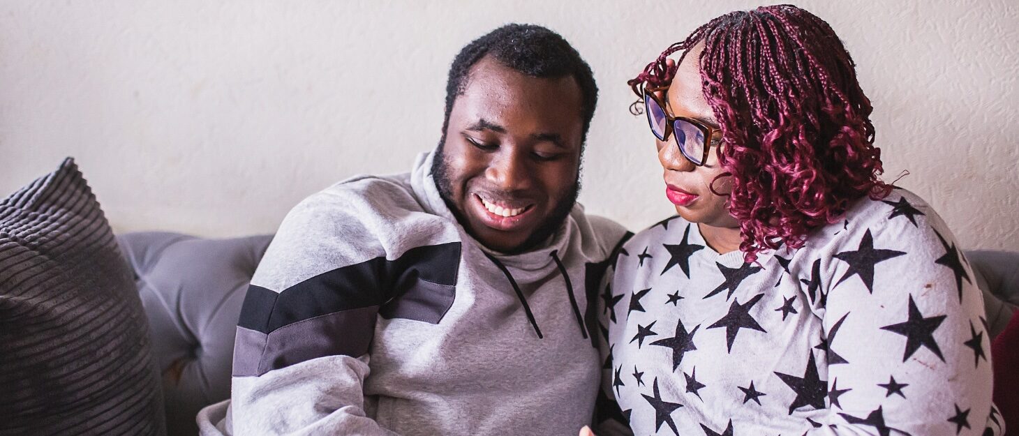 A young adult son smiling, sat on a sofa next to his mum wearing glasses and a starred t-shirt