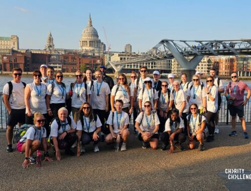 Large group of walkers in hiking equipment stood by Millennium Bridge in London, with the River Thames and St Paul's Cathedral in the background.