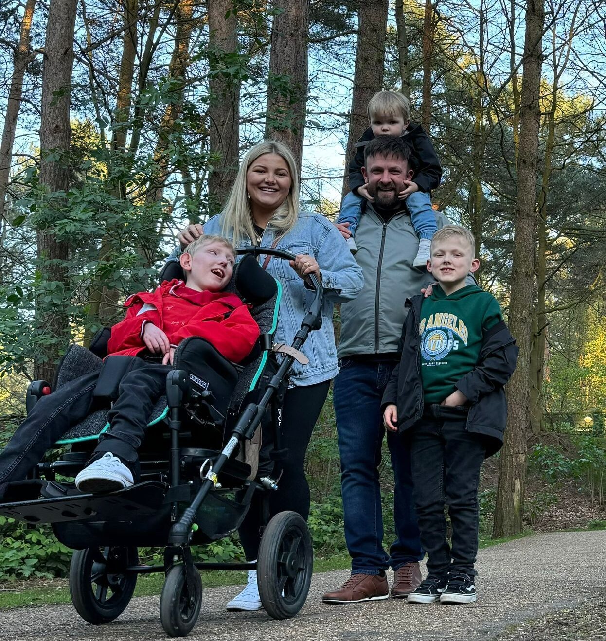 A family of 5 are smiling to the camera whilst on a woodland walk. Once child is in a wheelchair.