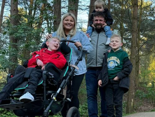 A family of 5 are smiling to the camera whilst on a woodland walk. Once child is in a wheelchair.