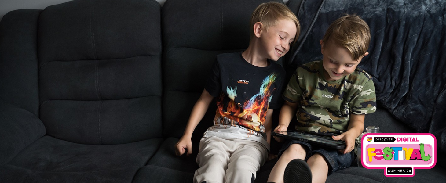 Two young brothers sit together on a sofa smiling whilst using a tablet.