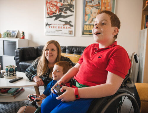 A young boy in a wheelchair plays on a games console with his younger brother, whilst mum watches on smiling.