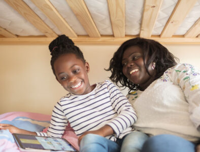 A young girl and her mum are laughing on a bottom bunk, playing on a tablet together.