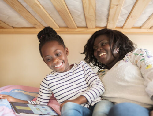 A young girl and her mum are laughing on a bottom bunk, playing on a tablet together.