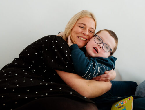 A mum and her young son hug on the sofa, smiling to the camera.