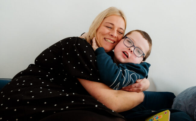 A mum and her young son hug on the sofa, smiling to the camera.