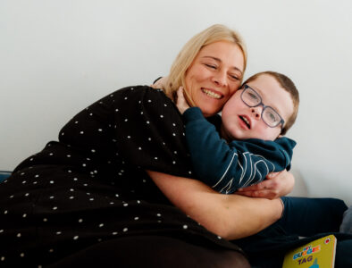 A mum and her young son hug on the sofa, smiling to the camera.