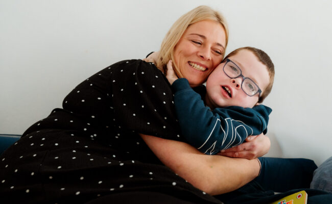 A mum and her young son hug on the sofa, smiling to the camera.