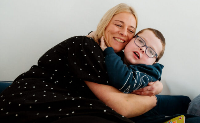 A mum and her young son hug on the sofa, smiling to the camera.