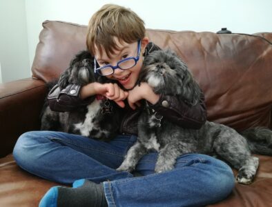 A young boy wearing glasses sits on a brown sofa and is cuddling two small dogs whilst smiling to the camera