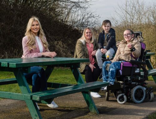 Woman sat on a picnic bench outside, next to her is a mum, her son and daughter in an electronic wheelchair