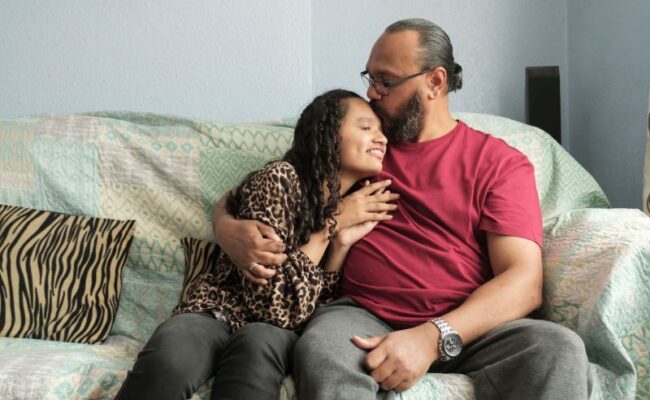 A young blind girl sitting on a sofa with her Dad kissing her forehead