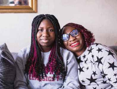 A teenage girl with long hair, and her mum seated next to here on a sofa smiling