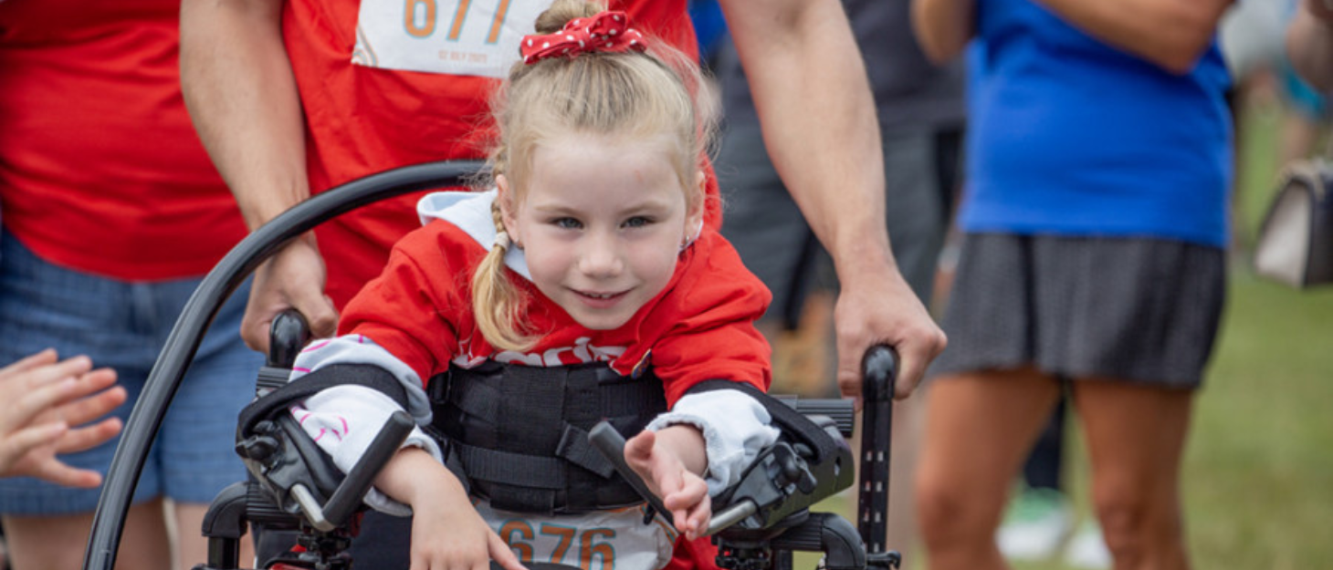 A young girl in a red top in a support frame with a guide pushing behind her