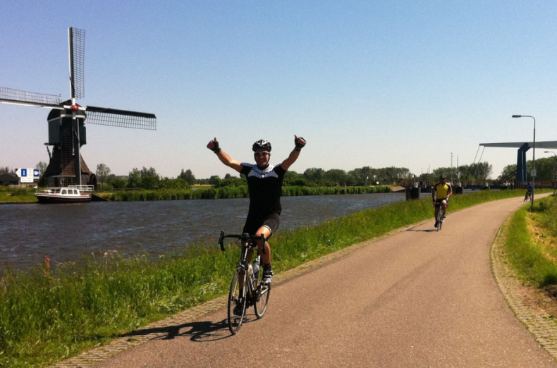 A man on a bicycle with his arms in the air celebrating completing the event. In the background is a river and a traditional Windmill