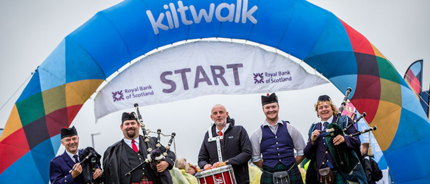 A large arch banner with 'Kiltwalk start' on it and in front are a group of people wearing kilts posing for a photo holding bagpipes and a drum