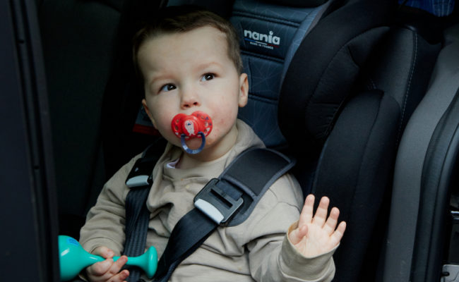 A little boy with a red dummy in his mouth is sat in a car seat waving