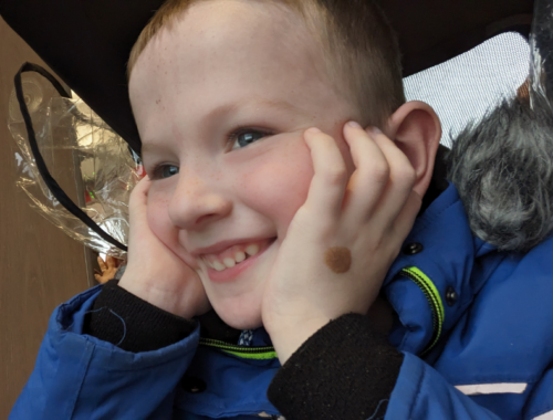 a little blond boy with blue eyes holds his face in his hands, looking far away, smiling