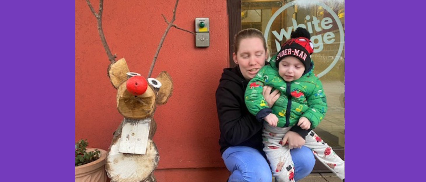 A mother holding her toddler, crouched outside next to a large wooden reindeer