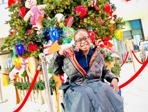 A young person in a wheelchair wearing glasses, poses in front of a large decorated Christmas tree.