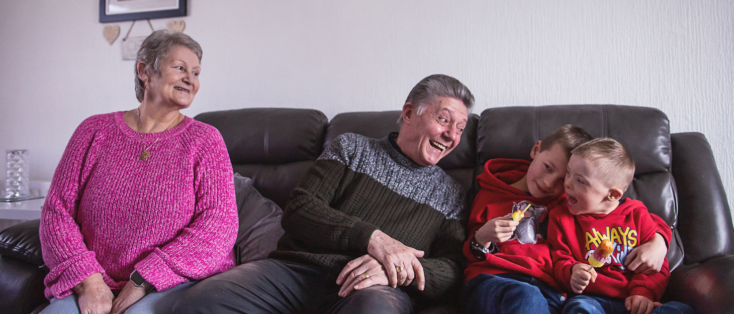 A gran and grandad sitting on a dark leather sofa looking over at their two young grandsons, who are holding ice lollies and smiling