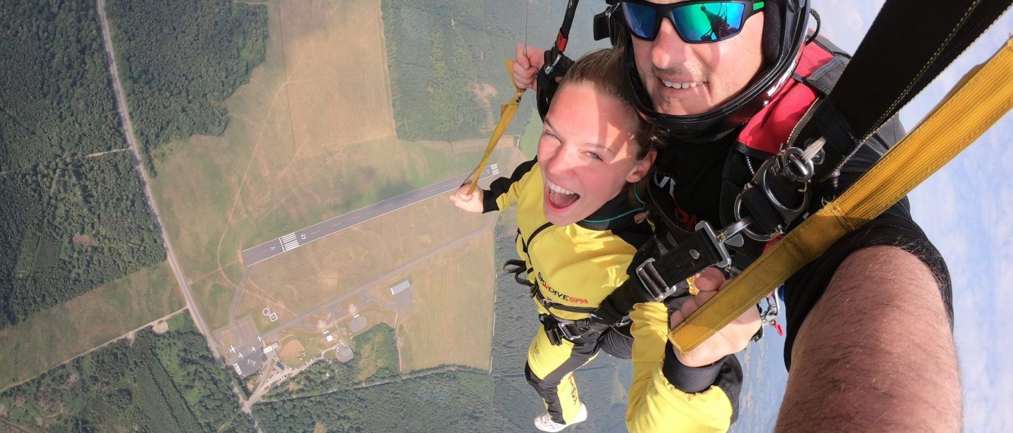 A selfie of a woman and man doing a tandem skydive with the airfield way below them