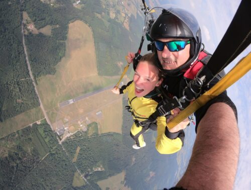 A selfie of a woman and man doing a tandem skydive with the airfield way below them