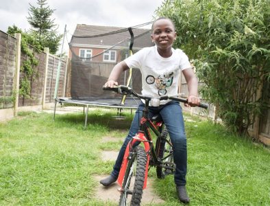 A boy riding a red bicycle in a garden with a trampoline behind him