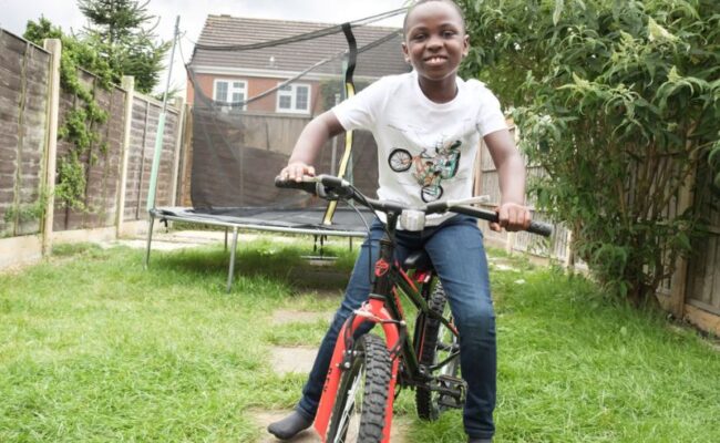 A boy riding a red bicycle in a garden with a trampoline behind him