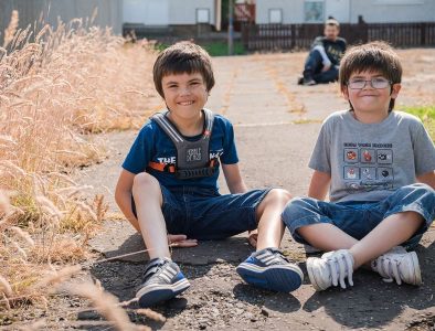 Two young boys wearing denim shorts are sat on the ground outside