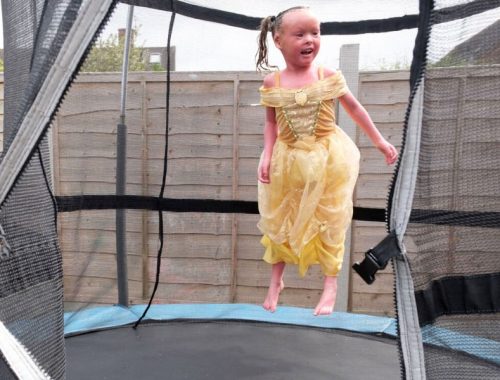 A little girl wearing a yellow princess dress bouncing on a trampoline outside