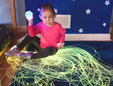 A young girl holding a light up ball sat in a blue room with sensory strand lights on the floor around her