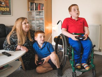 A boy in a wheelchair is using a games console with his younger brother and their mum sat with them