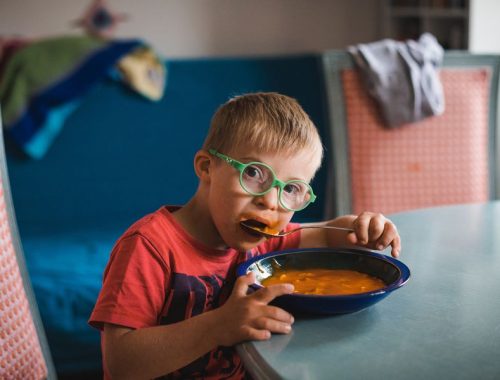 A little boy with green glasses eating a bowl of spaghetti with a spoon at a kitchen table