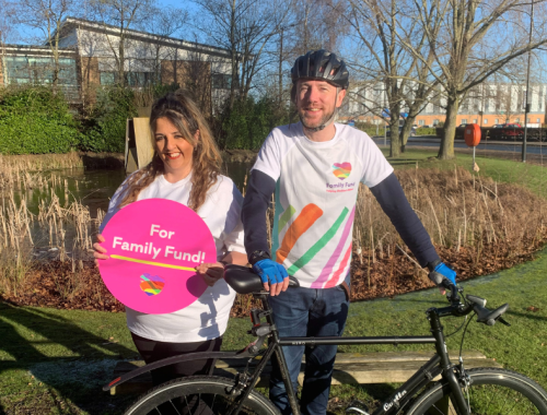 A woman and a man stand in front of a bicycle, smiling. The woman is holding up a pink Family Fund sign.