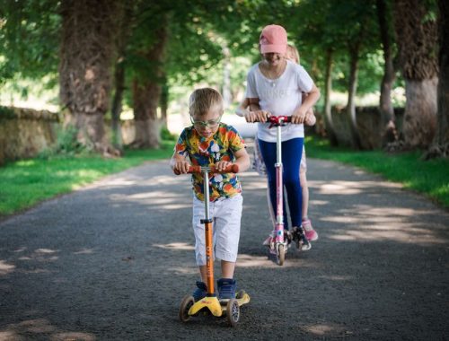 A young boy and his older sister riding scooters down a woodland path
