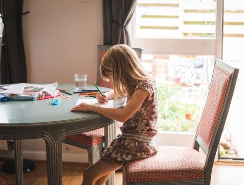 A girl with blonde hair drawing with pencils sat at a dining table