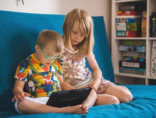 A little boy wearing green glasses looking at a tablet is sat next to his older sister