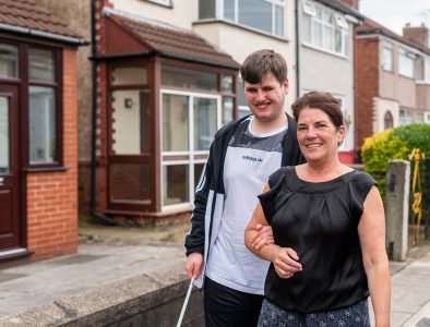 A mum with her teenage son who uses a walking cane, walking on the pavement of a street lined with houses