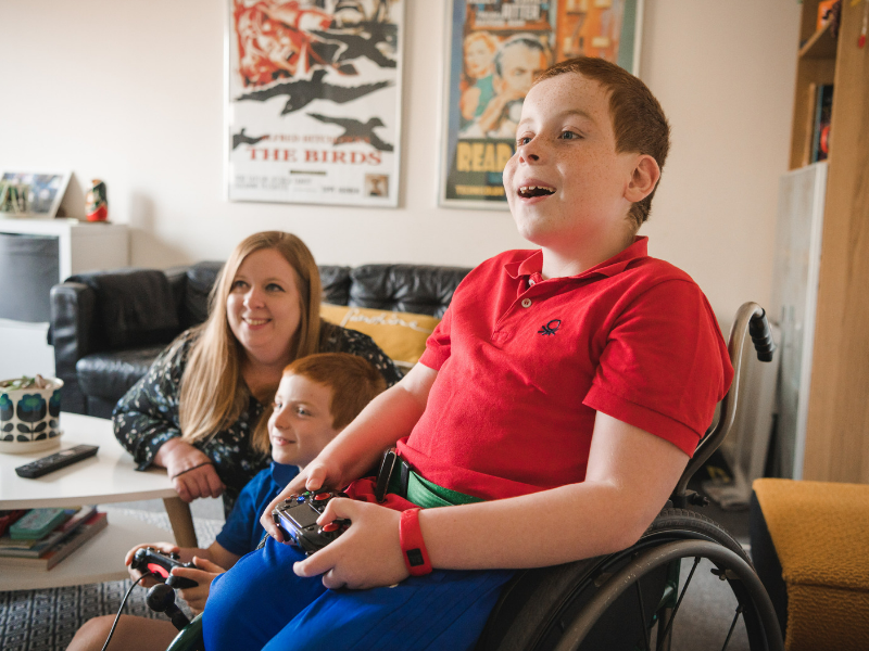 A boy in a red polo top sits in his wheelchair, smiling and playing on his games console. Behind him sit his mum and little brother.
