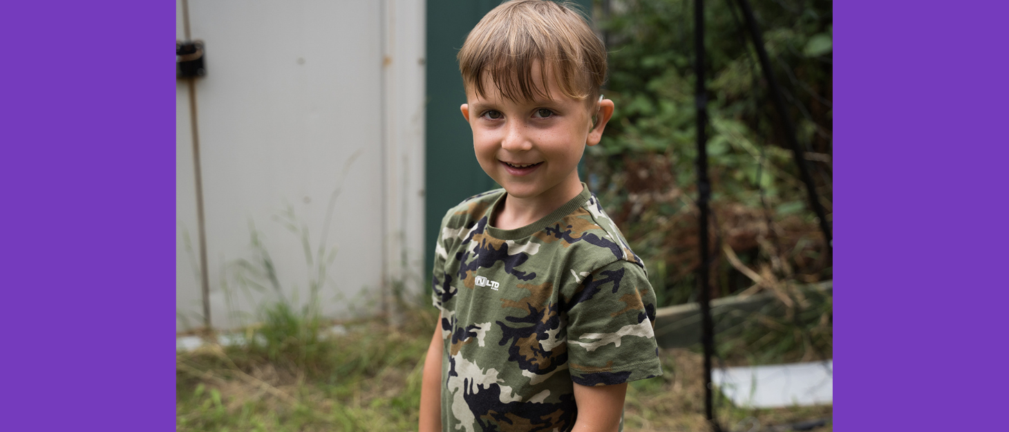 Boy standing outside in garden wearing a hearing aid smiling